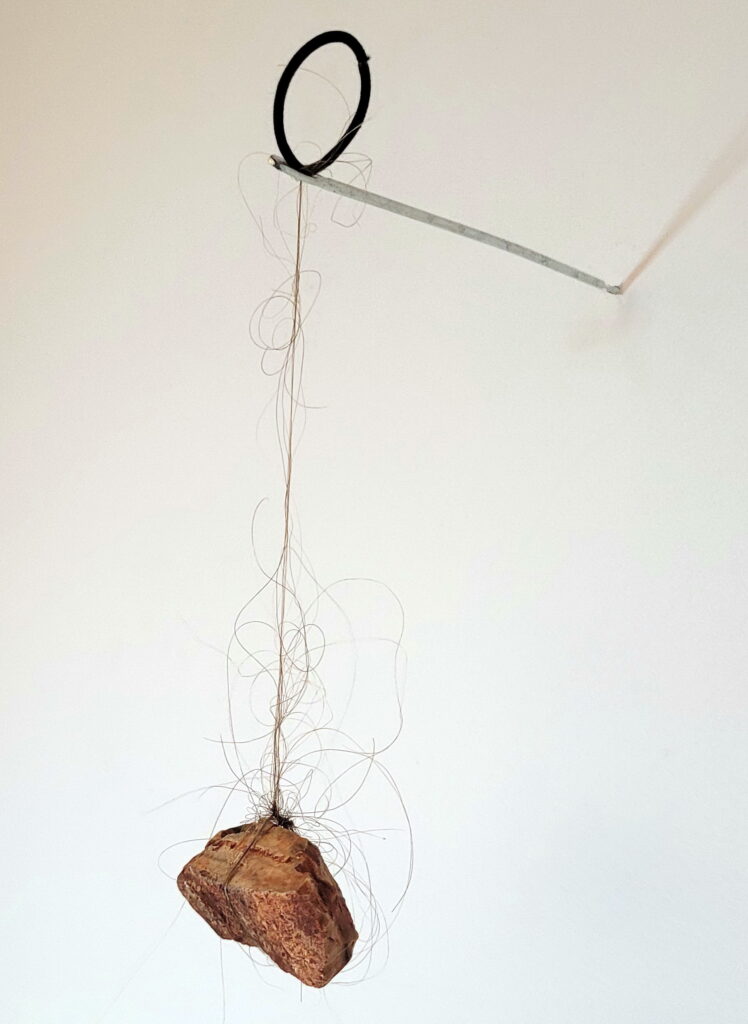 Fiando (series “O sustento”), 2023 Sculpture Artist’s hair, stone collected in Chapada Diamantina and hair elastic collected on the floor somewhere in the world, elastic band pin fixed to the wall 22 x 7 x 13cm 40g Single edition