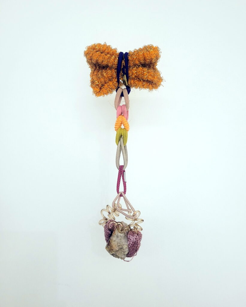 Inocência (series “O sustento”), 2023 Sculpture – Stone collected in Amsterdam – NL, 11 hair elastics collected on the ground in various places around the world, elastic band pin fixed to the wall 22 x 5 x 13cm 50g Single edition